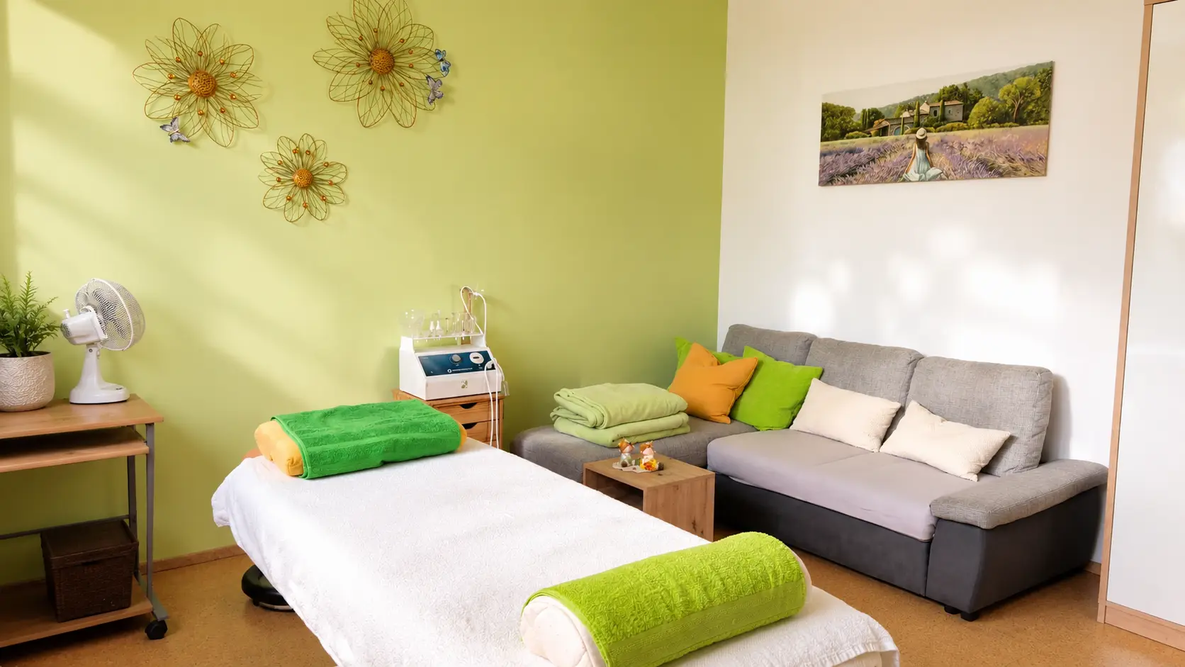 Heller Praxisraum von Hannelore Pritz mit Behandlungsliege, grünen Tüchern, Sofa und Blumendekor an der Wand